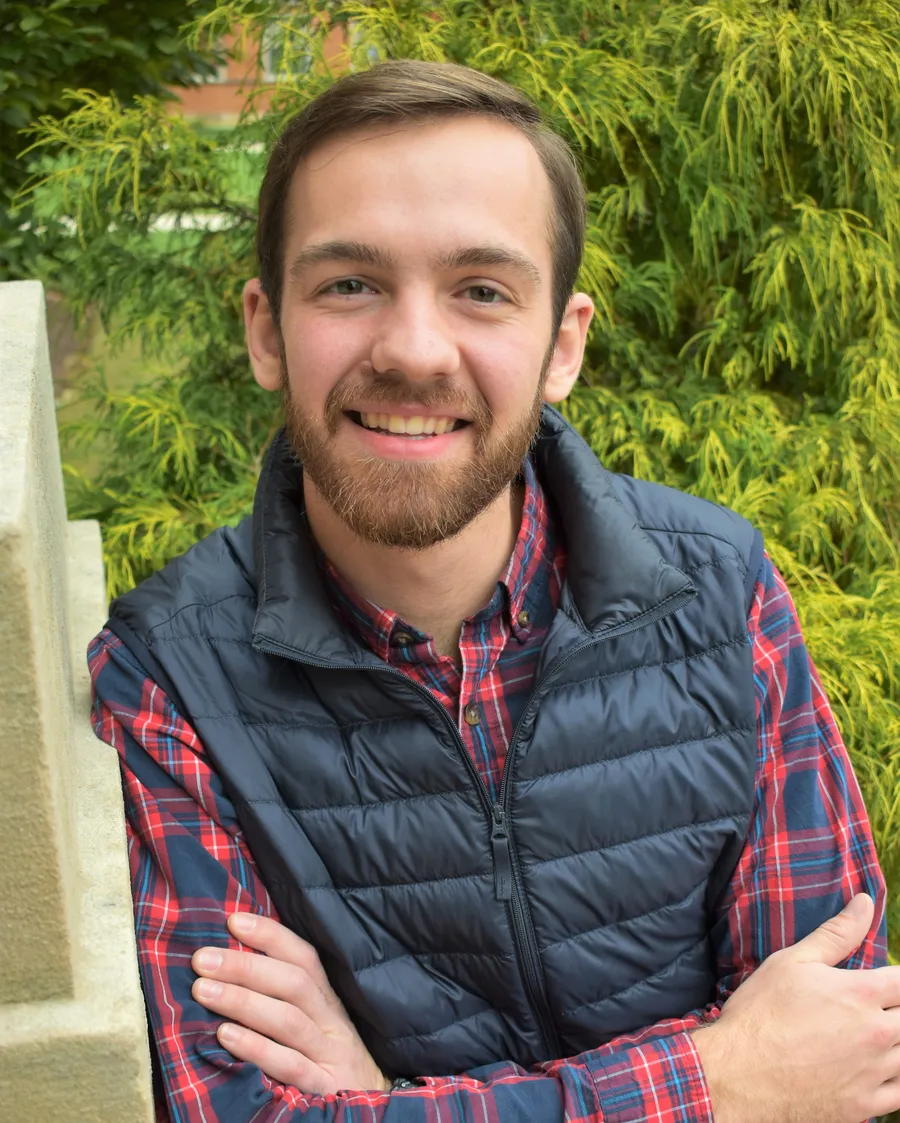 Portrait of Zachary Ratliff smiling, arms crossed, in front of evergreen shrubs